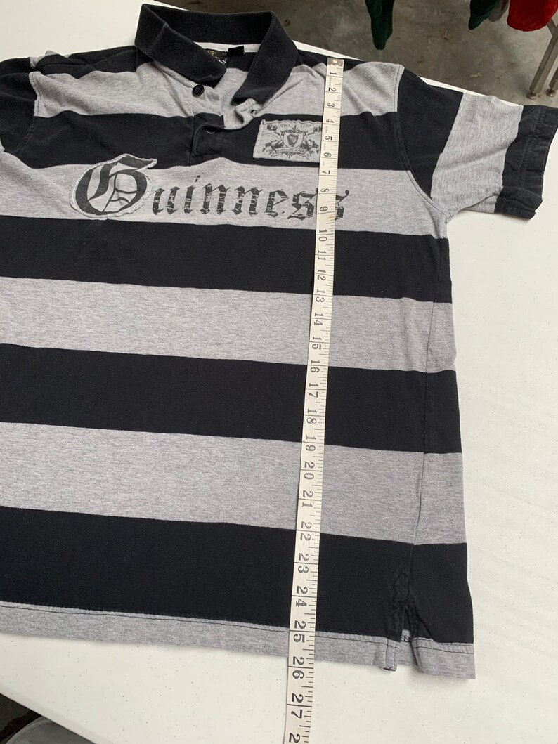 May include: A black and gray striped polo shirt with a Guinness logo on the front. The shirt has a button-down collar and short sleeves.