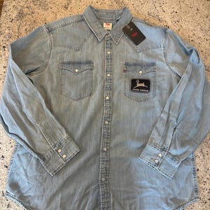 May include: Light blue denim shirt with two chest pockets and snap closures. Features a black patch with a deer graphic and the words "JOHN DEERE". Includes a Levi's tag.