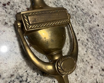 May include: A brass door knocker with a decorative design. The knocker has a rectangular plate with a rope-like border and a round knob at the bottom.