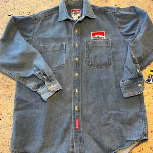 May include: A blue denim long-sleeve shirt with a red Marlboro logo patch on the left chest pocket. The shirt has a button-down collar and two front pockets.