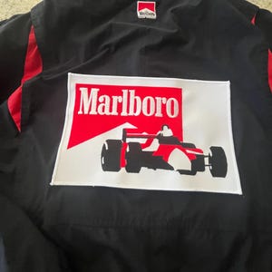 May include: A black jacket with red accents featuring a large rectangular patch on the back. The patch displays the red and white Marlboro logo above a black and white graphic of a race car. A smaller Marlboro logo is visible at the collar.