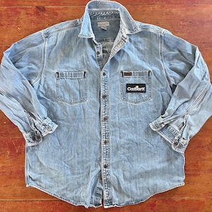 Puede incluir: Una camisa de mezclilla azul claro de manga larga con un parche de logotipo de Carhartt en el bolsillo izquierdo del pecho. La camisa tiene un frente abotonado y dos bolsillos en el pecho.