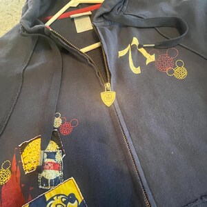 May include: A navy blue zip-up hoodie featuring a gold-toned shield-shaped zipper pull with a Mickey Mouse icon. The garment includes a graphic of a yellow Mickey Mouse head, red and yellow circular patterns, and a rectangular patch with a character.