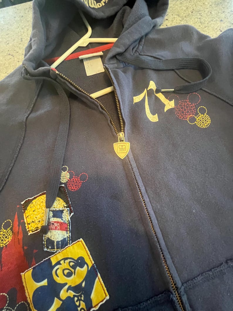 May include: A navy blue zip-up hoodie featuring a gold-toned shield-shaped zipper pull with a Mickey Mouse icon. The garment includes a graphic of a yellow Mickey Mouse head, red and yellow circular patterns, and a rectangular patch with a character.