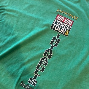 Op de afbeelding: Groen T-shirt met de tekst "PARTICIPANT" en "HOT ROD POWER TOUR 2011" in een gele doos. De tekst "INDIANAPOLIS" staat verticaal gestapeld in zwarte letters met de tekst "Indiana" eronder.