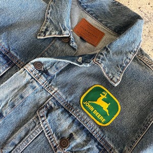 May include: A blue denim jacket with a brown leather patch that says "PREMIUM QUALITY LUCKY BRAND THE MARK OF GOOD STYLE" and a green and yellow John Deere patch with a deer on it.