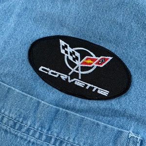 May include: A black oval patch with white stitching. The patch features a white Corvette logo with a red and yellow flag and the word "Corvette" in white lettering.