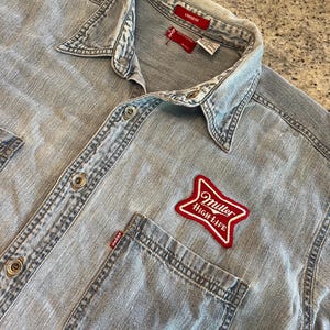 May include: Light blue denim shirt with a button-down front, two chest pockets, and a red Miller High Life patch. The shirt has a loose fit and a classic collar. The Levi's tag is visible.