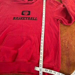 May include: Red hooded sweatshirt with a black basketball graphic and the word "BASKETBALL" in black lettering. The sweatshirt is measured with a tape measure.