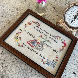 Puede incluir: Un sampler de punto de cruz enmarcado con un fondo blanco y bordado de colores. El sampler dice "No importa dónde sirva a mis invitados, parece que les gusta mi cocina." El sampler está enmarcado en un marco de madera oscura con un borde decorativo.