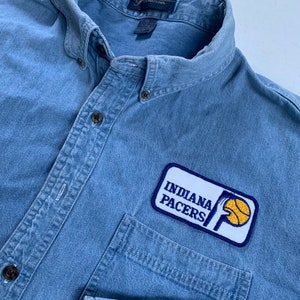 May include: A blue denim shirt with a patch on the left chest pocket. The patch is white with blue trim and features the Indiana Pacers logo, a basketball and the words "Indiana Pacers".