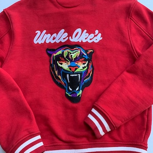 Puede incluir: Chaqueta de béisbol roja con rayas blancas en las mangas y el dobladillo inferior. La parte posterior de la chaqueta presenta un logotipo bordado en blanco que dice "Uncle Ike's" y un parche de cabeza de tigre colorido.