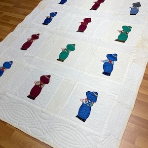 Könnte beinhalten: Eine weiße Steppdecke mit einem sich wiederholenden Muster aus gestickten Figuren. Die Figuren sind in blauer, roter und grüner Kleidung gekleidet. Die Steppdecke ist mit einem wellenförmigen Muster gesteppt.