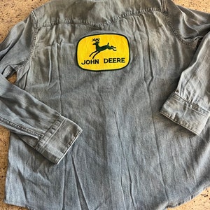 Pode incluir: Uma camisa de ganga azul claro com um remendo de logotipo John Deere amarelo e verde nas costas. O logotipo apresenta um veado saltando.