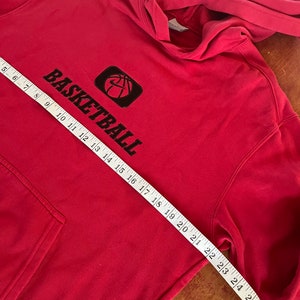 May include: Red hooded sweatshirt with a black basketball graphic and the word "BASKETBALL" printed on the front. The sweatshirt is measured with a tape measure.