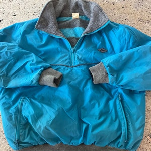 Puede incluir: Una chaqueta azul forrada de polar con cierre de cremallera y cuello, puños y cintura de canalé gris. La chaqueta tiene un pequeño logotipo en forma de diamante con la palabra "Pelle" en el pecho izquierdo.