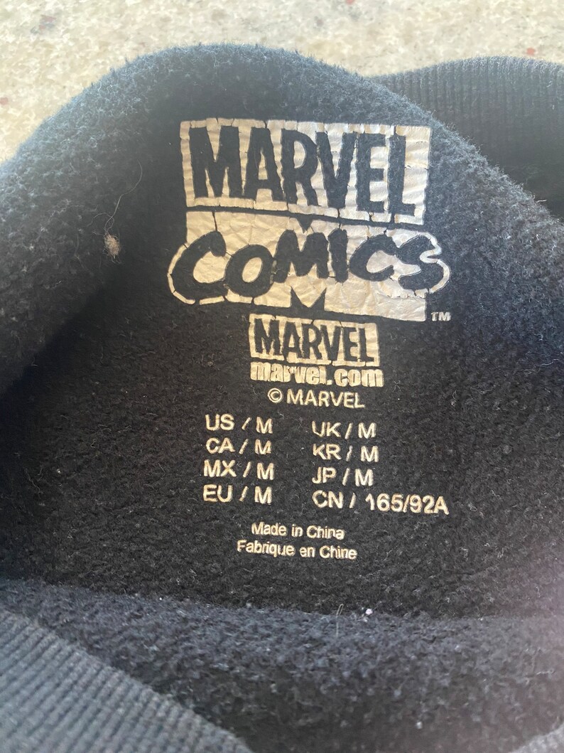 Puede incluir: Etiqueta negra con texto blanco que dice "MARVEL COMICS" y "MARVEL marvel.com" con un s&iacute;mbolo de copyright y un s&iacute;mbolo de marca registrada. La etiqueta tambi&eacute;n incluye informaci&oacute;n de tama&ntilde;o para EE. UU., Reino Unido, Canad&aacute;, Corea del Sur, M&eacute;xico, Jap&oacute;n y China.