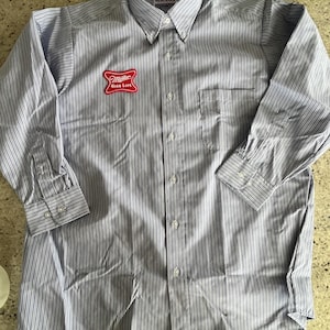 May include: A light blue and white striped button-down shirt with long sleeves. The shirt has a chest pocket and a red Miller High Life logo patch. The collar reads "HERCULES".