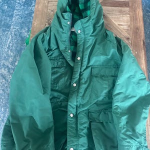 Puede incluir: Una chaqueta verde con capucha y forro de cuadros negros y verdes visible en la capucha. La chaqueta tiene cierre de cremallera, múltiples bolsillos y cierres de botón a presión blancos. La chaqueta está hecha de un material liso, posiblemente resistente al agua.