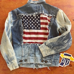 May include: A blue denim jacket with a distressed look, featuring a red, white, and blue American flag patch sewn onto the back. The jacket has a faded, bleached appearance and frayed edges.