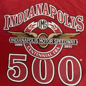 Może przedstawiać: Czerwona koszulka z napisem "Indianapolis" w stylizowanej czcionce, z "Indianapolis Motor Speedway" i "500" w kolorze białym i czerwonym. Koszulka upamiętnia Centennial Era, z datami 2009 i 2011.