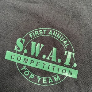 Puede incluir: Una camiseta negra con un gráfico circular verde que dice "First Annual S.W.A.T. Competition Top Team".
