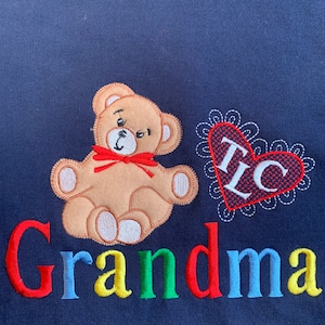 May include: A navy blue sweatshirt with a brown teddy bear, a red heart with the letters "I" and "C" inside, and the word "Grandma" embroidered in red, blue, green, and yellow.