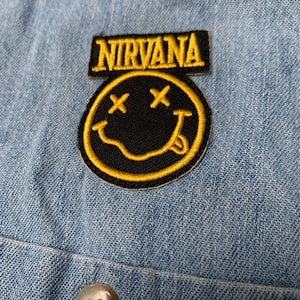 May include: A black and yellow embroidered patch with a smiley face and the word "NIRVANA" above it.