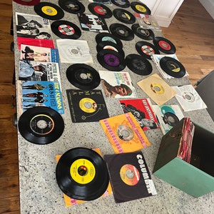May include: A collection of vintage 45 rpm vinyl records on a white countertop. The records are in various colors, including black, red, yellow, and blue. The records are arranged in a grid pattern. There is a green record storage box in the lower right corner of the image.