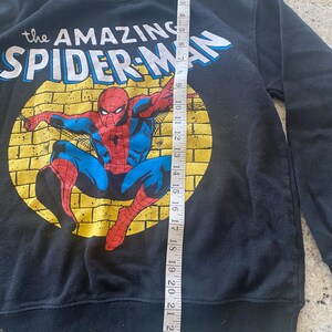 Puede incluir: Sudadera negra con un estampado gr&aacute;fico de Spider-Man en estilo de c&oacute;mic. El texto "the AMAZING SPIDER-MAN" est&aacute; impreso sobre la imagen.