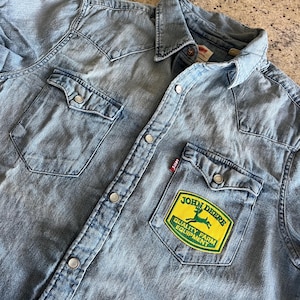 May include: A light blue denim shirt with a John Deere patch on the left pocket. The patch is yellow with green lettering and a green deer logo. The text on the patch reads "John Deere Quality Farm Equipment."