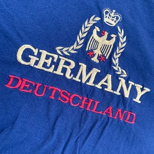 Puede incluir: Una camiseta azul con un águila bordada en blanco con una corona en la parte superior, rodeada de una corona de laurel. El texto "GERMANY" y "DEUTSCHLAND" está bordado en blanco y rojo.