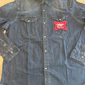 Może przedstawiać: Niebieska koszula dżinsowa z dwiema kieszeniami na piersi i czerwoną naszywką Miller High Life. Koszula ma klasyczny kołnierz, mankiety zapinane na guziki i karczek w stylu western. Wykonana z dżinsu w średnim odcieniu.