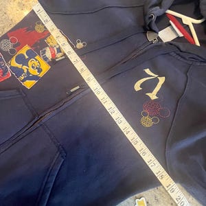 May include: A dark blue zip-up hoodie featuring a Mickey Mouse graphic and a gold embroidered logo. A white measuring tape is placed diagonally across the front, showing a length of approximately 22 inches from the shoulder to the bottom hem.