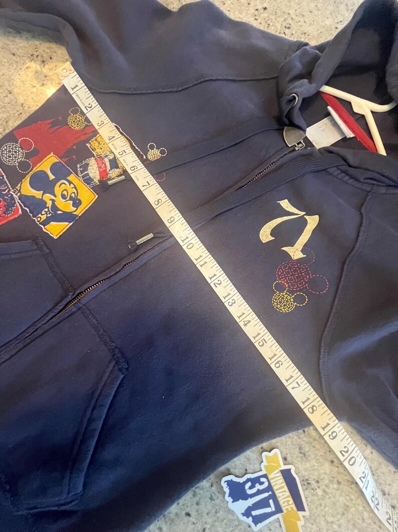 May include: A dark blue zip-up hoodie featuring a Mickey Mouse graphic and a gold embroidered logo. A white measuring tape is placed diagonally across the front, showing a length of approximately 22 inches from the shoulder to the bottom hem.
