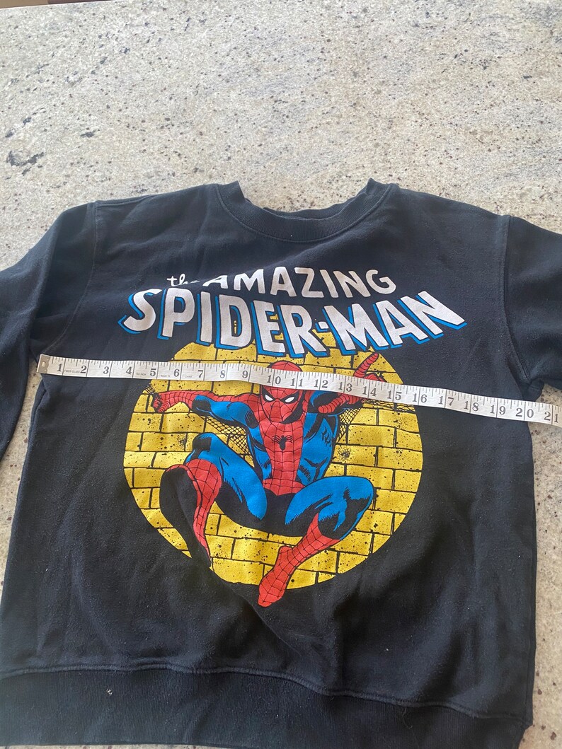 Puede incluir: Sudadera negra con un gr&aacute;fico de Spider-Man amarillo y rojo. El gr&aacute;fico dice "The Amazing Spider-Man".