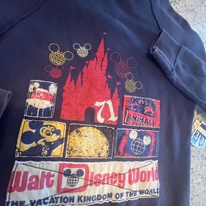 May include: A dark navy blue hoodie featuring a large graphic on the back. The design includes a red castle silhouette, various Mickey Mouse icons, and rectangular patches depicting Disney themes with the text Walt Disney World The Vacation Kingdom of the World.