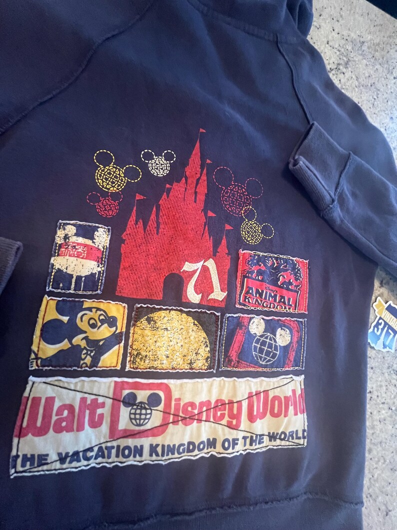 May include: A dark navy blue hoodie featuring a large graphic on the back. The design includes a red castle silhouette, various Mickey Mouse icons, and rectangular patches depicting Disney themes with the text Walt Disney World The Vacation Kingdom of the World.