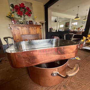 May include: A copper roasting pan with a stainless steel interior sits atop a copper pot with a single handle. The cookware is displayed on a wooden table, showcasing a classic kitchenware design.