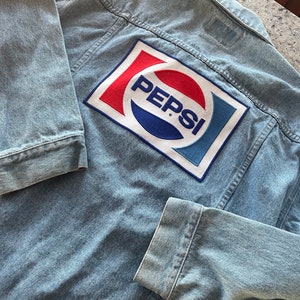 May include: A light blue denim jacket with a large red, white, and blue Pepsi logo patch on the back.