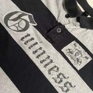 May include: Black and gray striped polo shirt with a black and white graphic of a basketball and the word "Timber" in a distressed font. The shirt has a small patch with a crest and the text "Est. 1759".