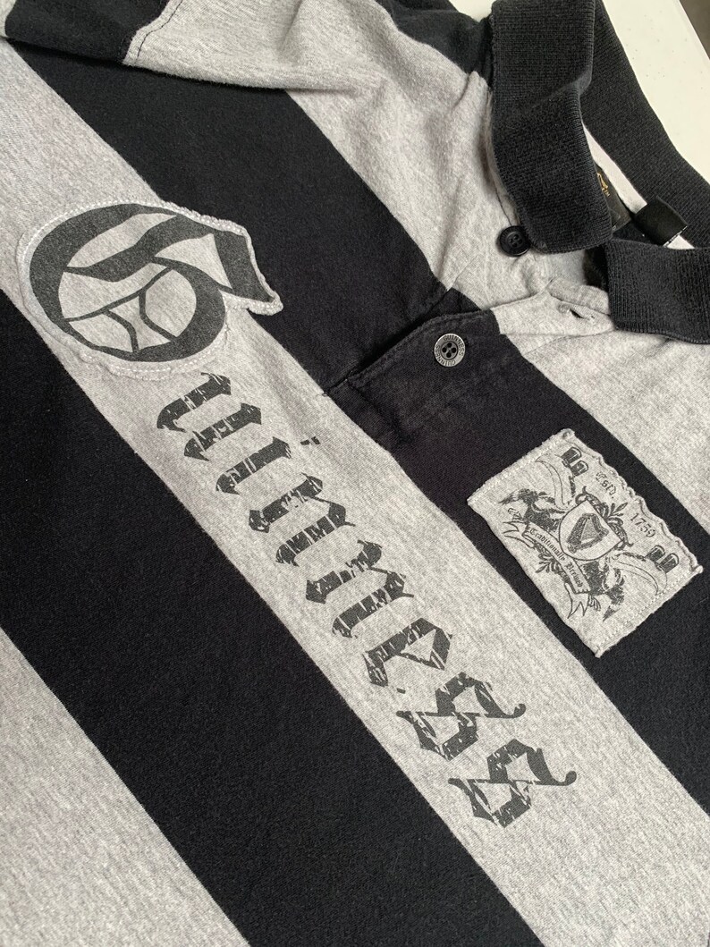 May include: Black and gray striped polo shirt with a black and white graphic of a basketball and the word "Timber" in a distressed font. The shirt has a small patch with a crest and the text "Est. 1759".