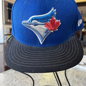 Peut inclure: Une casquette de baseball bleue et noire avec le logo des Blue Jays de Toronto représentant un oiseau stylisé avec une feuille d'érable rouge. La casquette est de la marque New Era.