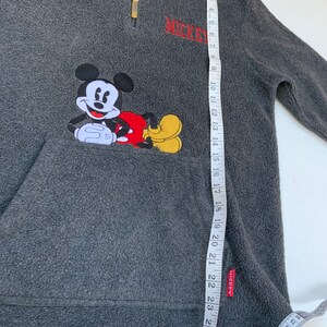 May include: Gray fleece pullover with a red and yellow Mickey Mouse applique. The pullover has a zipper and a pocket. The text "MICKEY" is embroidered on the front.