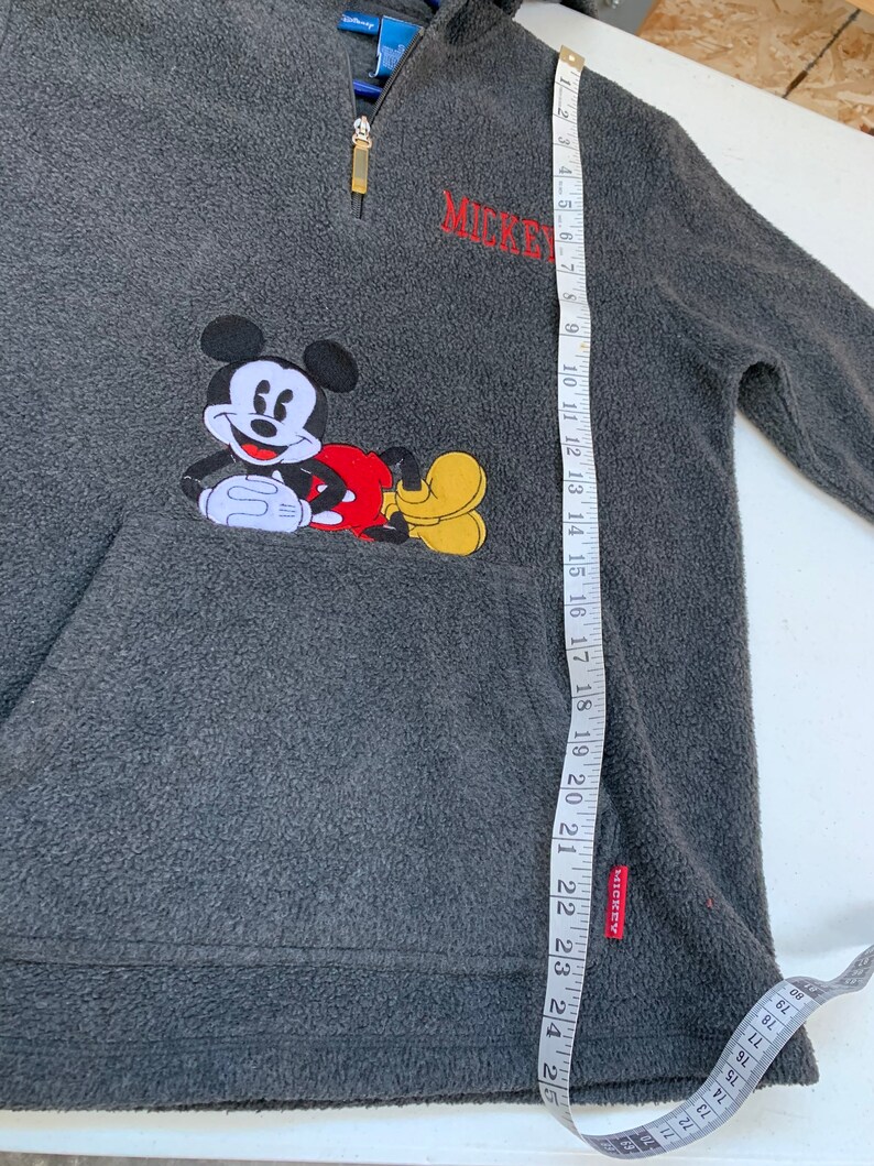 May include: Gray fleece pullover with a red and yellow Mickey Mouse applique. The pullover has a zipper and a pocket. The text "MICKEY" is embroidered on the front.