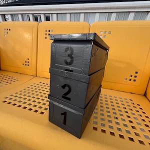 Op de afbeelding: Drie gestapelde, zwart geverfde dozen met de nummers 1, 2 en 3 staan op een gele bank. De dozen zijn rechthoekig en hebben grote, vette witte cijfers. De bank heeft een ruitvormig oppervlak en staat naast gele panelen.