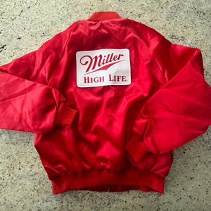 Może przedstawiać: Czerwona satynowa kurtka bomberka z białą i czerwoną naszywką z logo Miller High Life. Kurtka ma prążkowany kołnierz, mankiety i dół. Kurtka jest w żywym czerwonym kolorze.