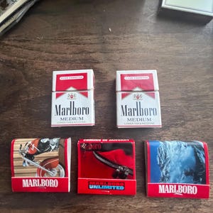 May include: Two packs of Marlboro Medium cigarettes and three Marlboro matchboxes are displayed. The matchboxes feature images of a motorcycle, a zipper, and a winter scene. The packs are white with red accents.