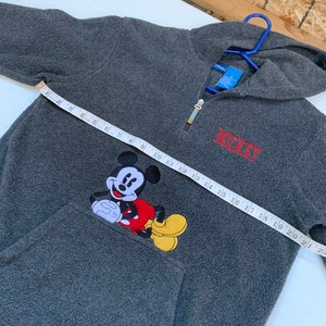 May include: Gray fleece pullover with a red Mickey Mouse graphic and the word "MICKEY" embroidered in red on the front. The zipper is gold and the hoodie has a drawstring.
