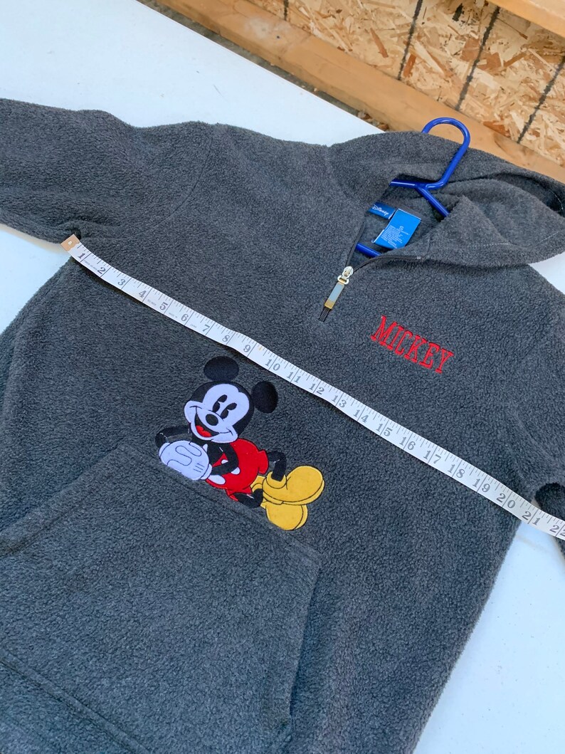 May include: Gray fleece pullover with a red Mickey Mouse graphic and the word "MICKEY" embroidered in red on the front. The zipper is gold and the hoodie has a drawstring.
